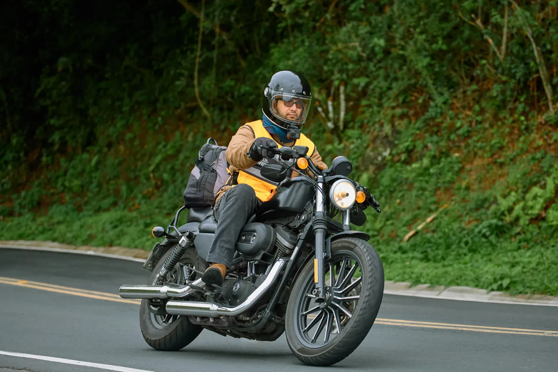 A rider on a Harley-Davidson Sportster taking a bend on a mountain road, in a black helmet and an orange vest